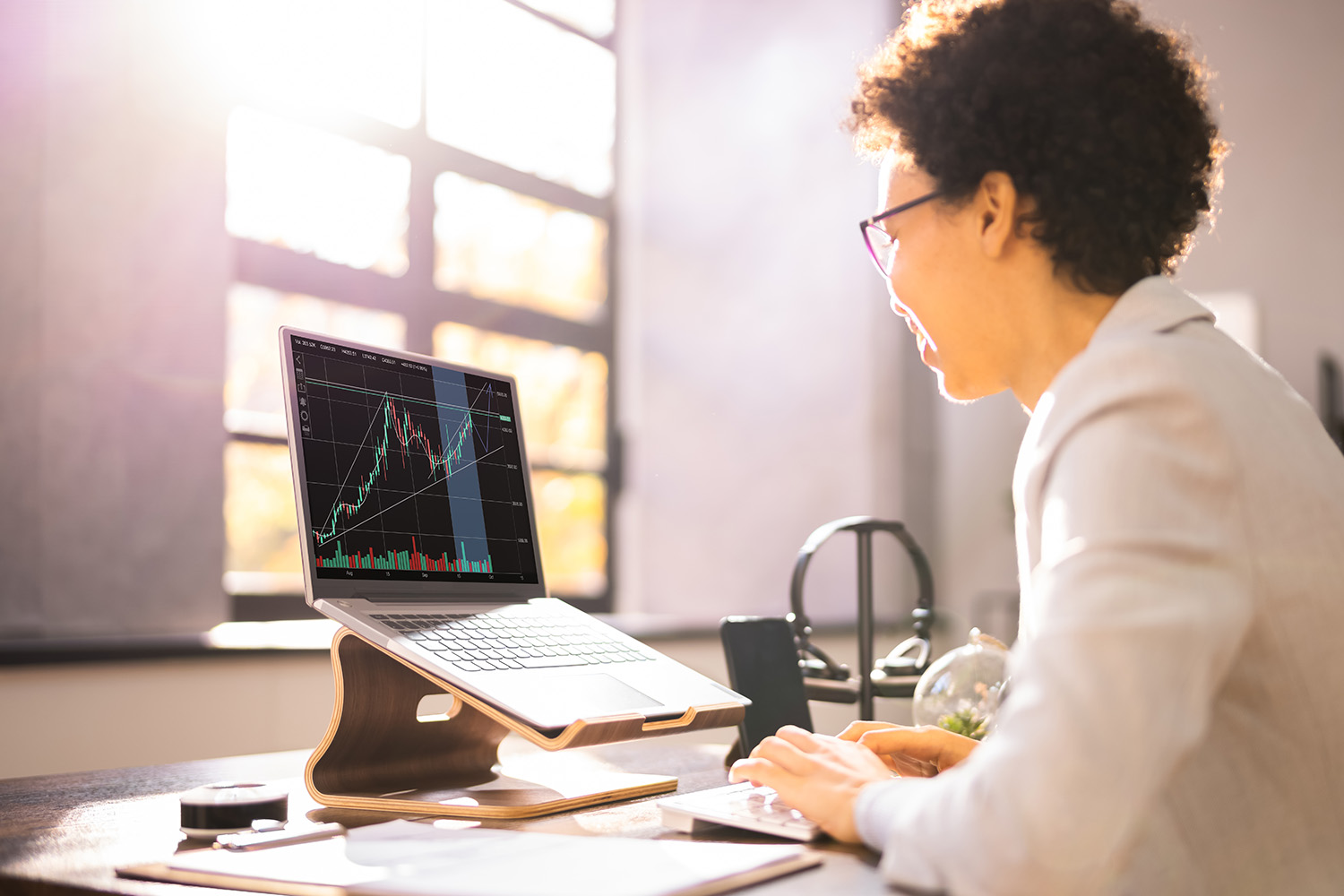 A woman in an office with light shining through the window, on a laptop with the stock market showing.